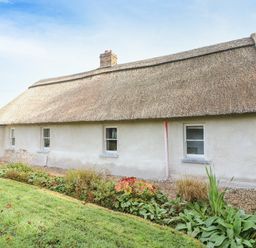 A thatched roof cottage with white walls and small windows surrounded by grass and plants at New Thatch Farm in Scarteen near Knocklong County Limerick