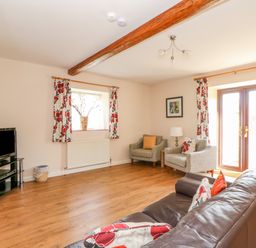 A living room with a brown sofa, two armchairs, a TV on a stand, floral curtains, and wooden floor at The Granary Cottage in Gayton near King's Lynn