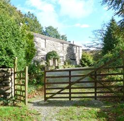 A stone cottage behind a wooden gate and stone walls surrounded by trees and greenery at High Kiln Bank Cottage in Seathwaite Duddon Valley