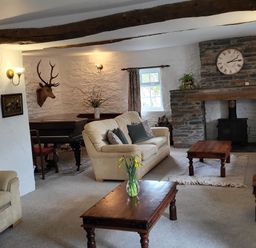 A living room with beige sofas wooden coffee tables a stone fireplace with a clock a mounted deer head and a piano at High Kiln Bank Cottage in Seathwaite Duddon Valley