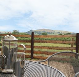 A metal outdoor table with glass and metal coffee pot and cups next to a wooden fence with hills in the background at Bicton Lodge in Clun