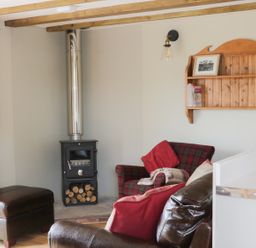 A living room with a wood stove a plaid armchair a leather ottoman and a wooden wall shelf at Bicton Lodge in Clun