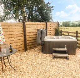 An outdoor area with a hot tub wooden steps a metal table with a bottle and glasses and a closed umbrella at Bicton Lodge in Clun