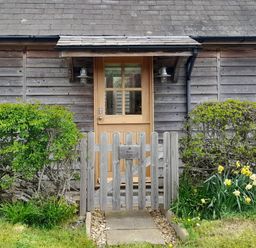 A wooden front door with a small pitched roof and gate surrounded by bushes and flowers at Bicton Lodge in Clun