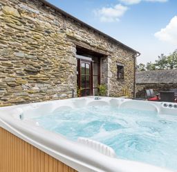 An outdoor hot tub on a stone patio next to a stone building with wooden doors and outdoor dining furniture with red cushions at Hill Side Barn in Pennington near Ulverston