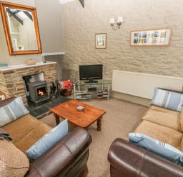 A living room with two sofas a wooden coffee table a fireplace with a wood stove a television on a glass stand and framed pictures on a stone wall at Foxglove Cottage in Little Haven near Haverfordwest