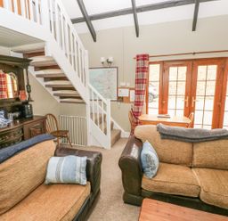 A living room with two brown sofas a wooden dining table and chairs a staircase with white railing and glass doors with red plaid curtains at Foxglove Cottage Little Haven near Haverfordwest