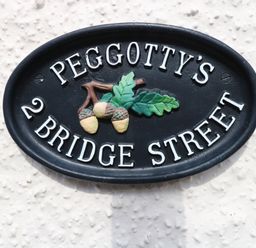 A black oval house sign with white letters reading Peggotty's 2 Bridge Street with a design of three acorns and two oak leaves at Peggotty's in Kelsale near Saxmundham