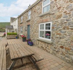 A stone house exterior with white framed windows and a wooden picnic table on a brick patio with flower pots at Hillbrook House in Nolton Haven