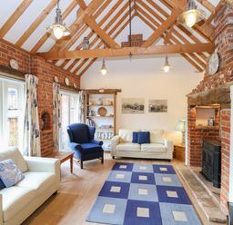 A living room with two white sofas a blue armchair a patterned rug a brick fireplace and exposed wooden beams at Stable Cottage in Necton near Swaffham