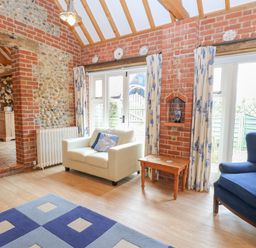 A sitting room with brick walls a white armchair a blue armchair a wooden side table and a blue patterned rug at Stable Cottage in Necton near Swaffham