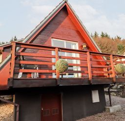 A red wooden chalet with a balcony and hanging plants in a wooded area at Larchfield Chalet 2 in Heights of Achterneed near Strathpeffer
