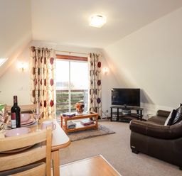 A living room with a brown leather sofa a TV on a stand a coffee table and a dining table set with dishes and a bottle of wine at Larchfield Chalet 2 Heights of Achterneed near Strathpeffer