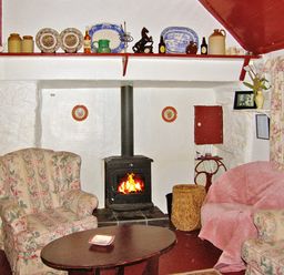 A living room with a wood stove fireplace two floral armchairs a round wooden table and shelves with decorative plates at Carthy's Cottage Kilbrien near Dungarvan County Waterford