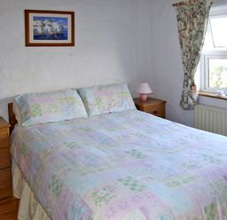 A bedroom with a bed between two wooden bedside tables with lamps and a window with floral curtains at Delia's Cottage in Ballinrobe County Mayo