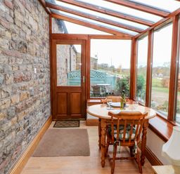 A small dining area with a round wooden table and two chairs next to a glass door and windows with a stone wall at The Stable in Milton near Brampton