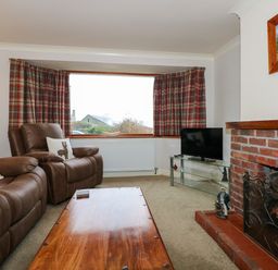 A living room with brown leather chairs a wooden coffee table a brick fireplace and a window with plaid curtains at Brynteg in Llwyngwril