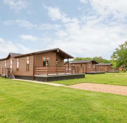 Brown wooden lodges with porches on green lawns under a cloudy sky at Callow Lodge 15 Beaconsfield Farm Holiday Park near Shrewsbury