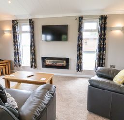 A living room with leather sofas a wooden coffee table a wall-mounted television and curtains at Callow Lodge 15 Beaconsfield Farm Holiday Park near Shrewsbury
