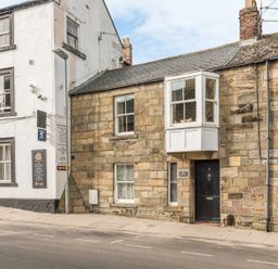 A street view showing a stone house with a black door and bay window next to a white building with window signs at Jackson Cottage in Alnmouth