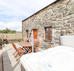An outdoor patio with a wooden table and chairs a hot tub a charcoal grill and a stone wall at Isallt Llanfair near Harlech