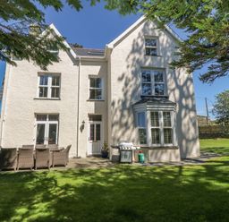 A white two-story house with multiple windows patio furniture and a barbecue on a lawn at Tyddyn Goronwy in Talybont