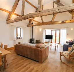 A living room with wooden beams a brown sofa a blue armchair a wooden dining table and chairs and a wood stove at Hill Farm Cottage in Kerrys Gate near Abbey Dore