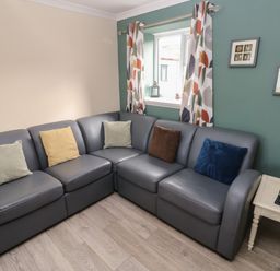 A corner sofa with cushions next to a window with patterned curtains and a small table with a remote control at Barn Cottage in Laugharne