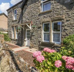 The stone exterior of a house with a white door wooden bench and pink flowers at Riverside View in Cark in Cartmel