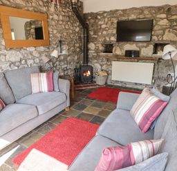 A living room with two gray sofas with striped cushions a wood burning stove a wall-mounted TV and stone walls at The Coach House in Wolfscastle