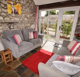A sitting room with two grey sofas, a red rug, a wooden side table, stone walls, and glass doors at The Coach House in Wolfscastle