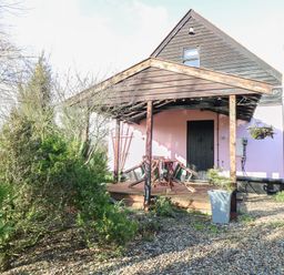 A wooden porch with a table and chairs in front of a house surrounded by trees and gravel at The Haybarn in Necton near Swaffham