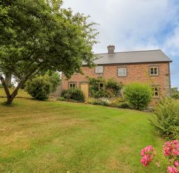 A brick house with multiple windows surrounded by a garden with trees bushes and flowers at Byrdir Cottage in Rhayader