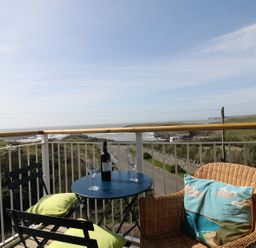 A balcony with a round table holding a bottle of wine and two wine glasses two chairs one with green cushions and one wicker chair with a cushion overlooking the sea at Seascape in Trearddur Bay