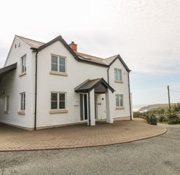 A two story white house with multiple windows and a small porch surrounded by a paved driveway at Seascape in Trearddur Bay