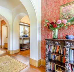 A hallway with archways a carved wooden bookshelf with books and flowers a coat rack and rugs on wooden floor at Plas Gwynfryn in Llanbedr