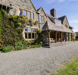 A stone house with ivy on the walls a covered porch with wooden chairs and a gravel driveway at Plas Gwynfryn in Llanbedr