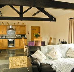 An open plan kitchen and living area with wooden cabinets a dining table with chairs and a black sofa with cushions and a throw at The Byre in Llanddewi Skirrid near Abergavenny