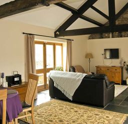 A living room with a dining table near wooden doors and a black sofa under exposed ceiling beams at The Byre in Llanddewi Skirrid near Abergavenny