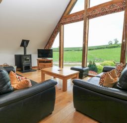 A living room with black leather sofas wooden coffee table wood stove and large windows overlooking a green field at Shepherd's Barn Between Milborne St Andrew and Winterborne Whitechurch