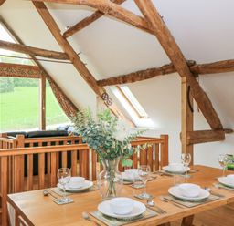 A dining table set with bowls glasses and cutlery near wooden beams and windows showing a green field at Shepherd's Barn Between Milborne St Andrew and Winterborne Whitechurch