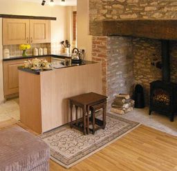 A kitchen area with wooden cabinets and a kettle beside a stone fireplace with a wood stove and firewood at Candy Cottage Thornford near Sherborne