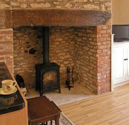 A room with a wood-burning stove set in a brick and stone fireplace a wooden table cups on a countertop and a television on a white cabinet at Candy Cottage Thornford near Sherborne