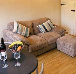 A living room with a brown sofa, striped cushions, ottoman, round table with wine bottle, glasses and fruit bowl at Candy Cottage Thornford near Sherborne