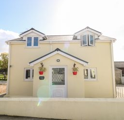 A two-story pale yellow house with white framed windows and a white front door with two red flower pots on either side of the entrance at Saddlers Cottage Berllandeg Farm