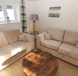 A living room with two beige sofas a round wooden coffee table a tripod floor lamp a painting of trees on the wall and a window with green plaid curtains at Saddlers Cottage Berllandeg Farm Clunderwen near Narberth
