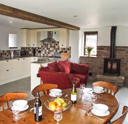 A kitchen with cream cabinets next to a dining table set with glasses bottles and bowls and a red sofa near a stone fireplace at Bushmills in Kerrys Gate near Abbey Dore