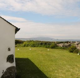 A garden with grass and shrubs overlooking a body of water with a mountain in the distance at Ty Fferm Bodlasan in Llanfachraeth near Valley