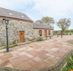 A stone building with wooden doors and windows on a paved courtyard with lamp posts and flower beds at Daffodil Cottage in Caeathro near Caernarfon