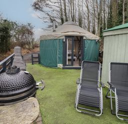 An outdoor area with two black lounge chairs a round yurt with green covering a black barbecue grill and a wooden shed at The Lakeside Yurt in Beckford